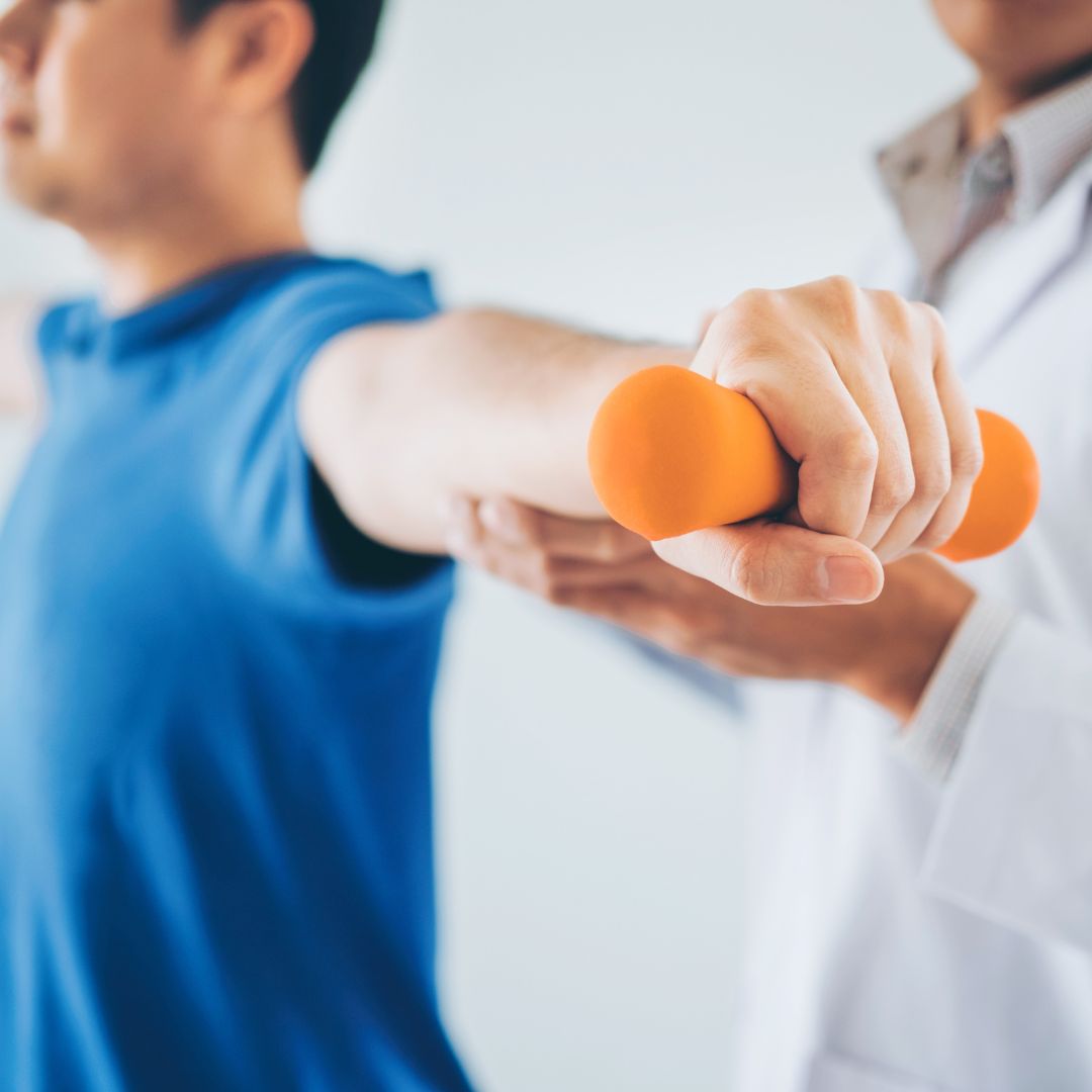Close-up of a patient holding an orange dumbbell while a physical therapist in a white coat guides their extended arm.
